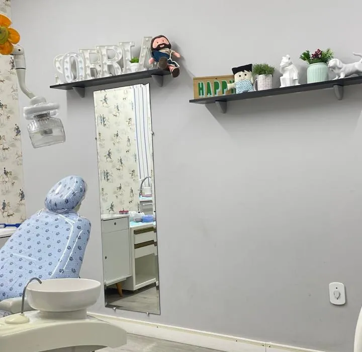 Parede cinza com um espelho alto e prateleiras com as letras SORRIA e bichinhos, ao lado da cadeira odontológica.