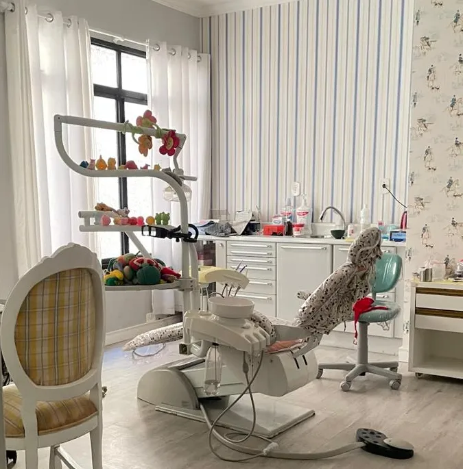 Sala de atendimento com parede listrada de azul, cadeira odontológica e cadeira de encosto xadrez amarelo.